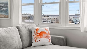 A grey sofa with a white cushion featuring a crab design, positioned in front of a window with a view of buildings and power lines. - Golden Shores Penthouse (Ocean City)