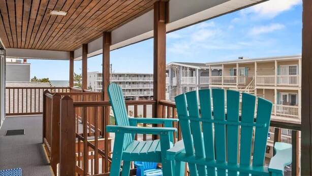 A covered outdoor area with wooden railings, two turquoise Adirondack chairs, and a view of adjacent buildings. - Golden Shores Penthouse (Ocean City)