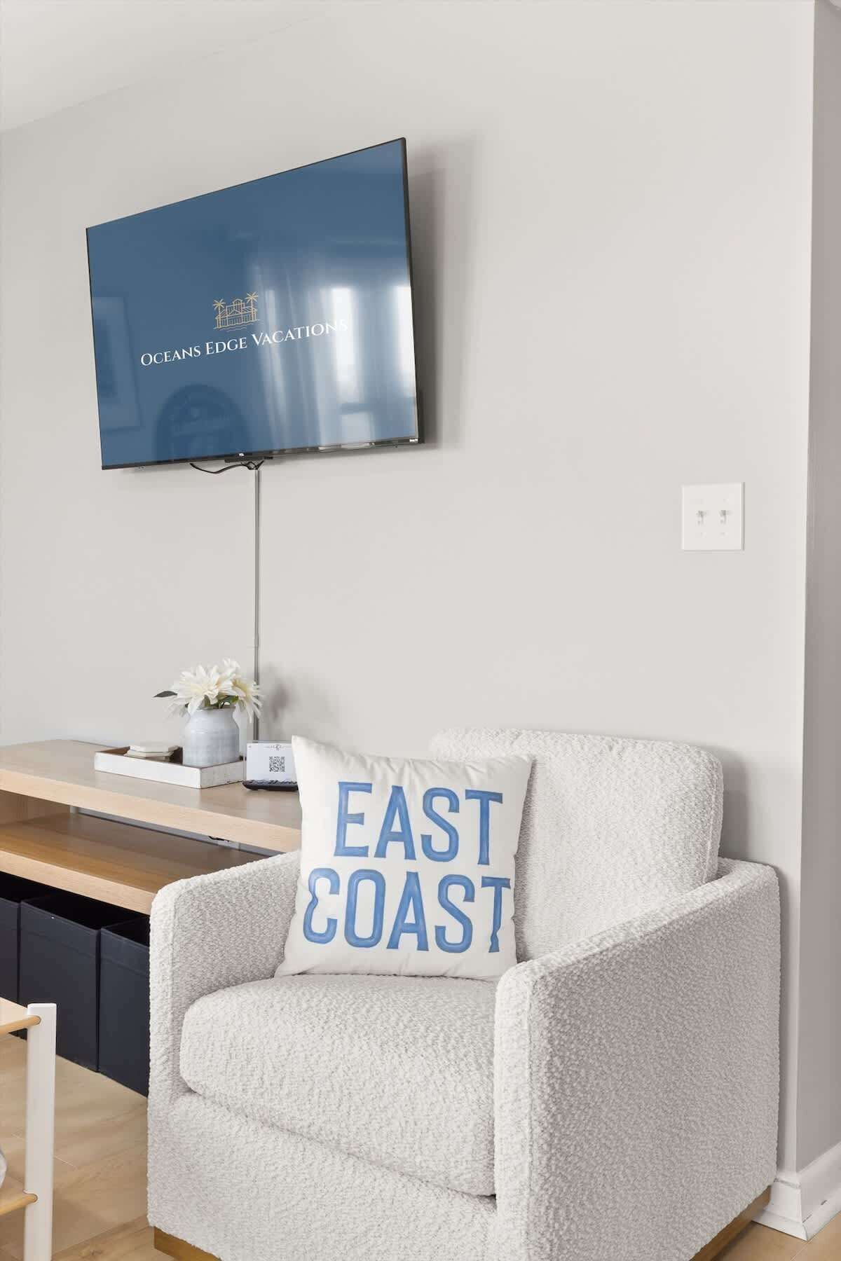 A living room with a white armchair, a flat-screen TV mounted on the wall, and a cushion with "EAST COAST" text.