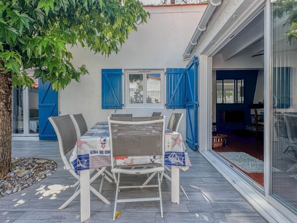 An outdoor patio with a table and chairs, a tree, and a building with blue shutters. - 4-room house near the sea with WIFI and pets allowed (La Palmyre)