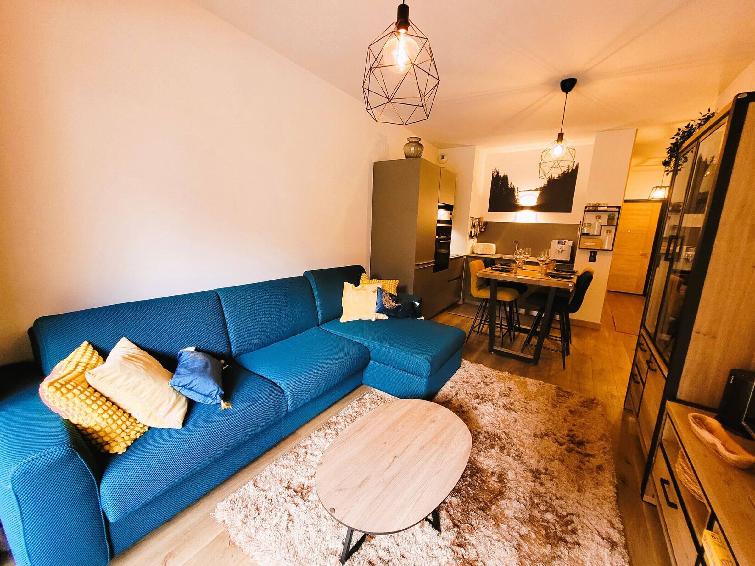 Un salón moderno con un sofá azul, una mesa de centro redonda de madera y una zona de cocina al fondo.