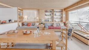 Un salon chaleureux et bien éclairé, avec un plafond en bois, un coin repas comprenant une table et des chaises en bois, et une grande fenêtre donnant sur un paysage enneigé.