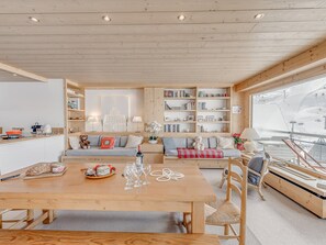 Uma sala de estar acolhedora e bem iluminada, com teto de madeira, área de refeições com mesa e cadeiras de madeira, e uma grande janela com vista para uma paisagem coberta de neve.