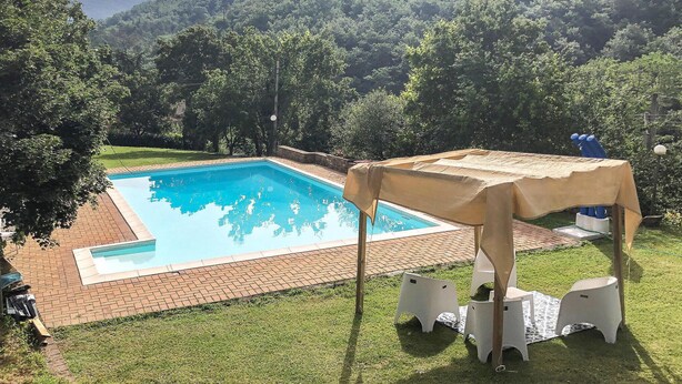 A rectangular swimming pool with a clear blue water surface, surrounded by a brick deck. There are white plastic chairs and a table with a beige cover. The setting is outdoors with a grassy area and trees in the background. - Lovely home in Arezzo with WiFi (Arezzo)