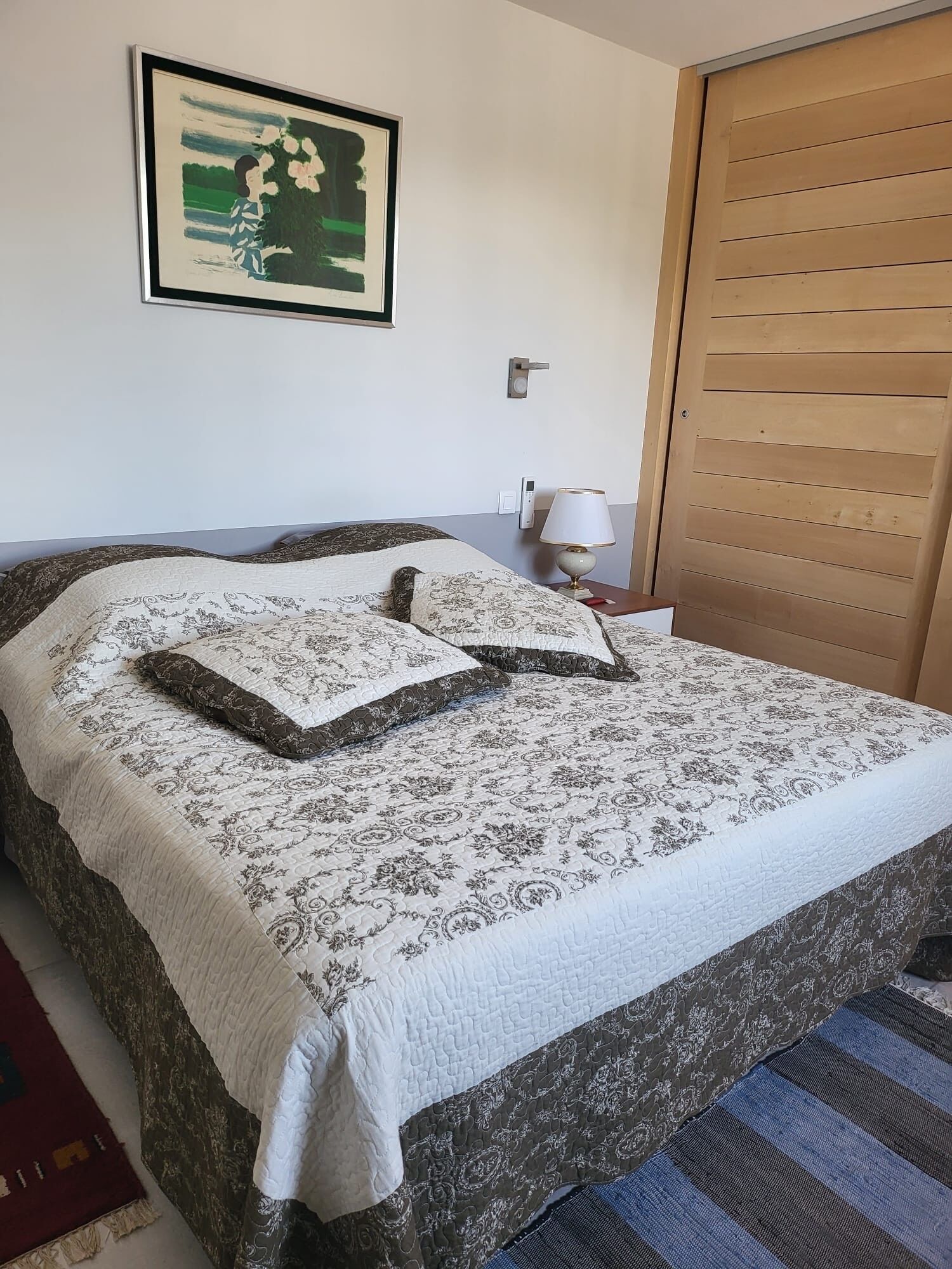 Un lit bien fait, avec une couvre-lit à motifs et des oreillers, une lampe de chevet, un tableau encadré au mur et une armoire en bois.