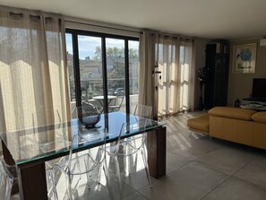 Un saló modern amb una taula de dinar de vidre, cadires de plàstic transparent, un sofà groc i grans finestres amb cortines.