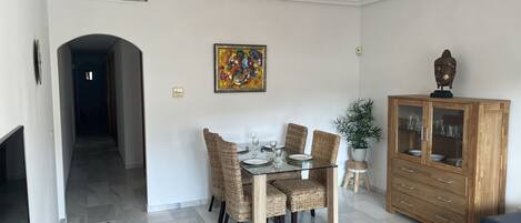Un comedor con una mesa dispuesta para cuatro personas, rodeada de sillas de mimbre, un mueble con puertas de cristal y una estatua de Buda encima.
