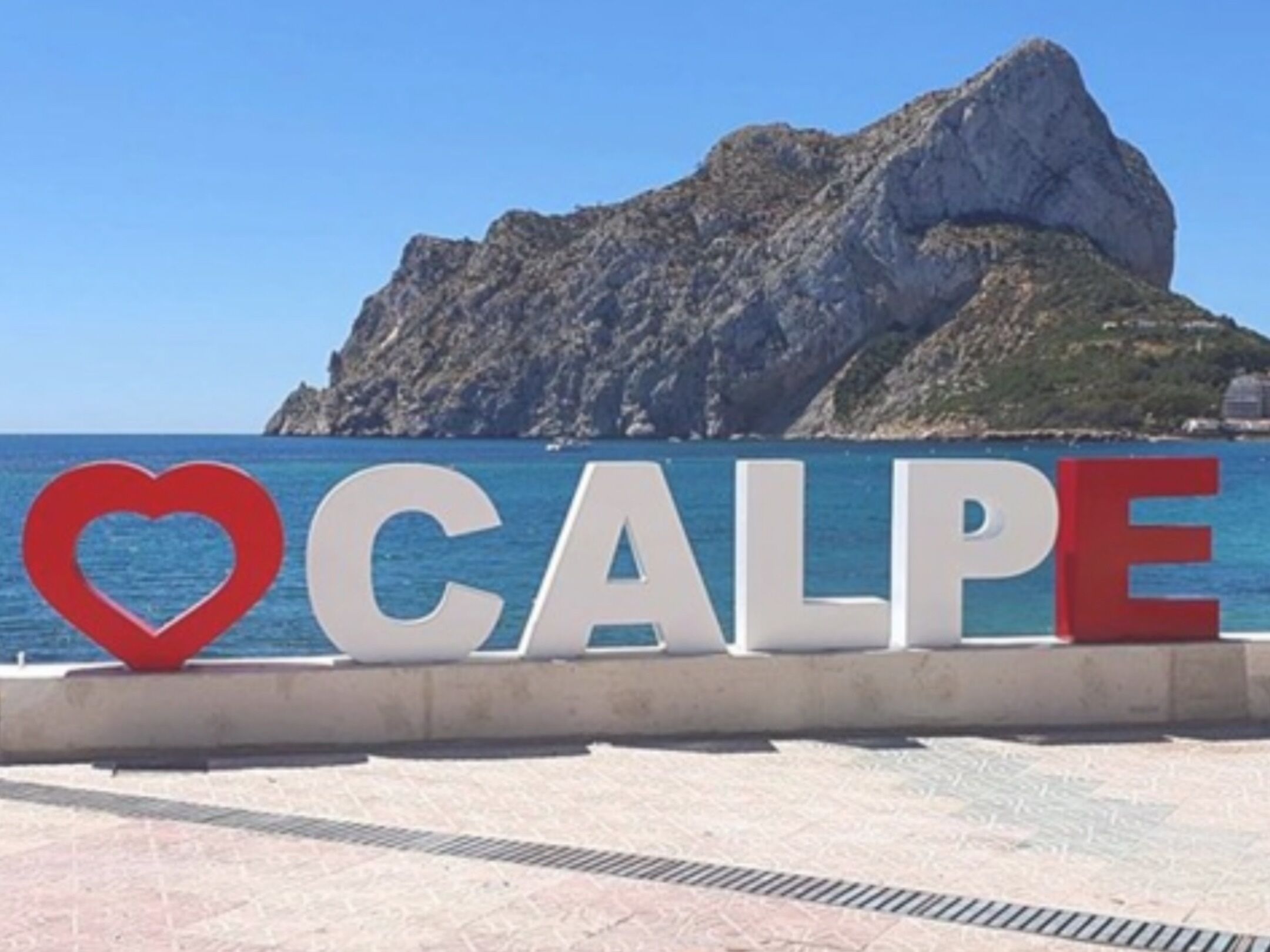 Ein Schild mit dem Wort 'CALPE' und einem Herzsymbol vor einer felsigen Küstenlinie und blauem Meer.