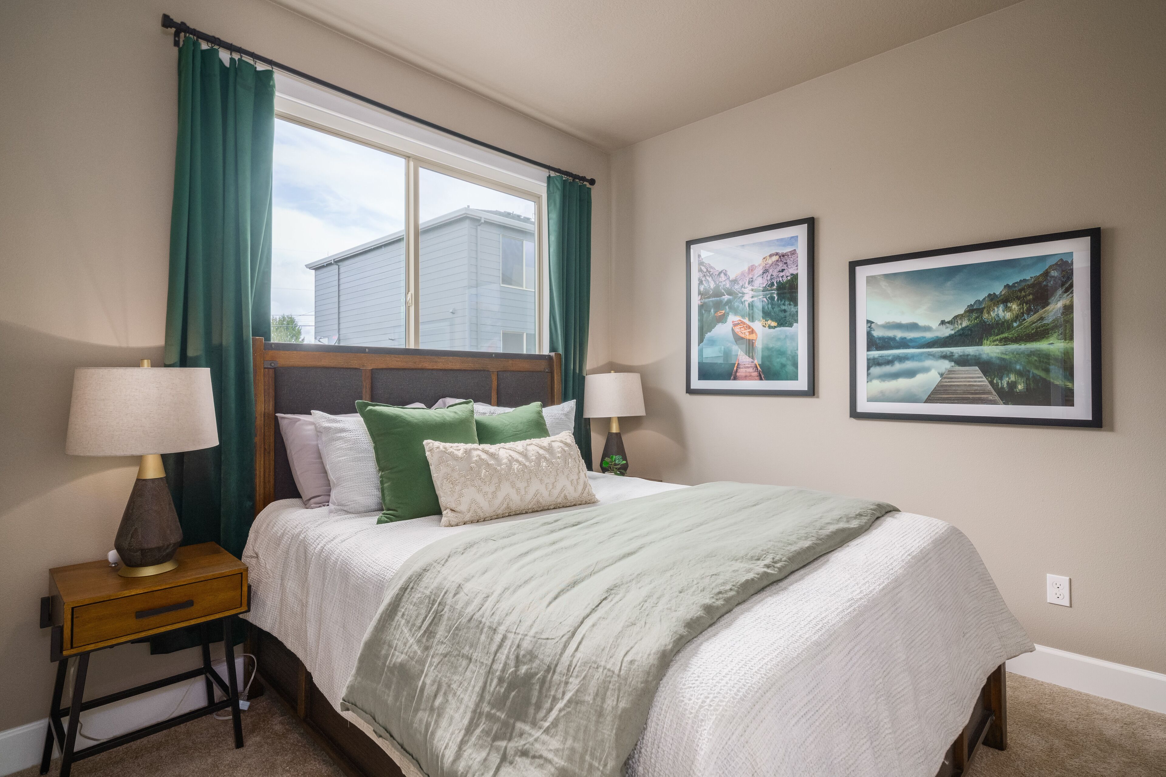 A bedroom with a bed, two bedside lamps, a window with curtains, and two framed pictures on the wall.
