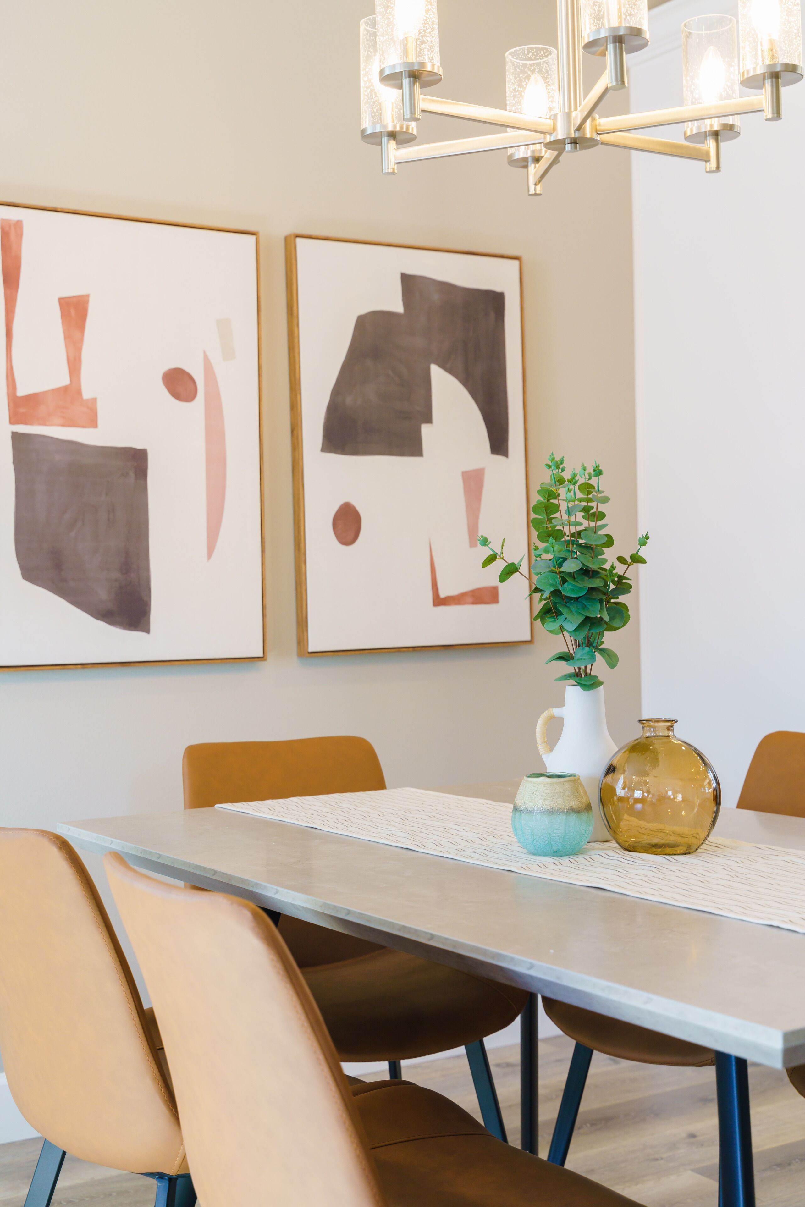 A dining area with a table, chairs, and a chandelier. There are two framed abstract paintings on the wall.