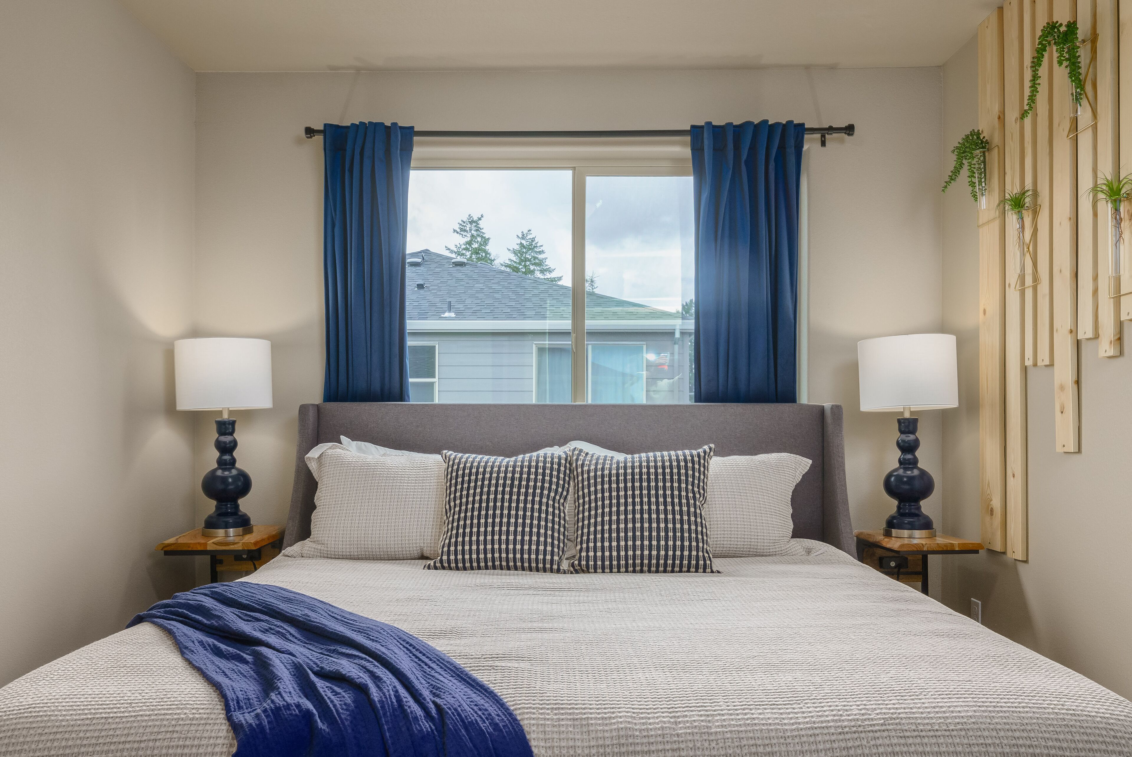 Una camera da letto con un letto grande, due comodini con lampade, una finestra con tende azzurre e vista su una casa.