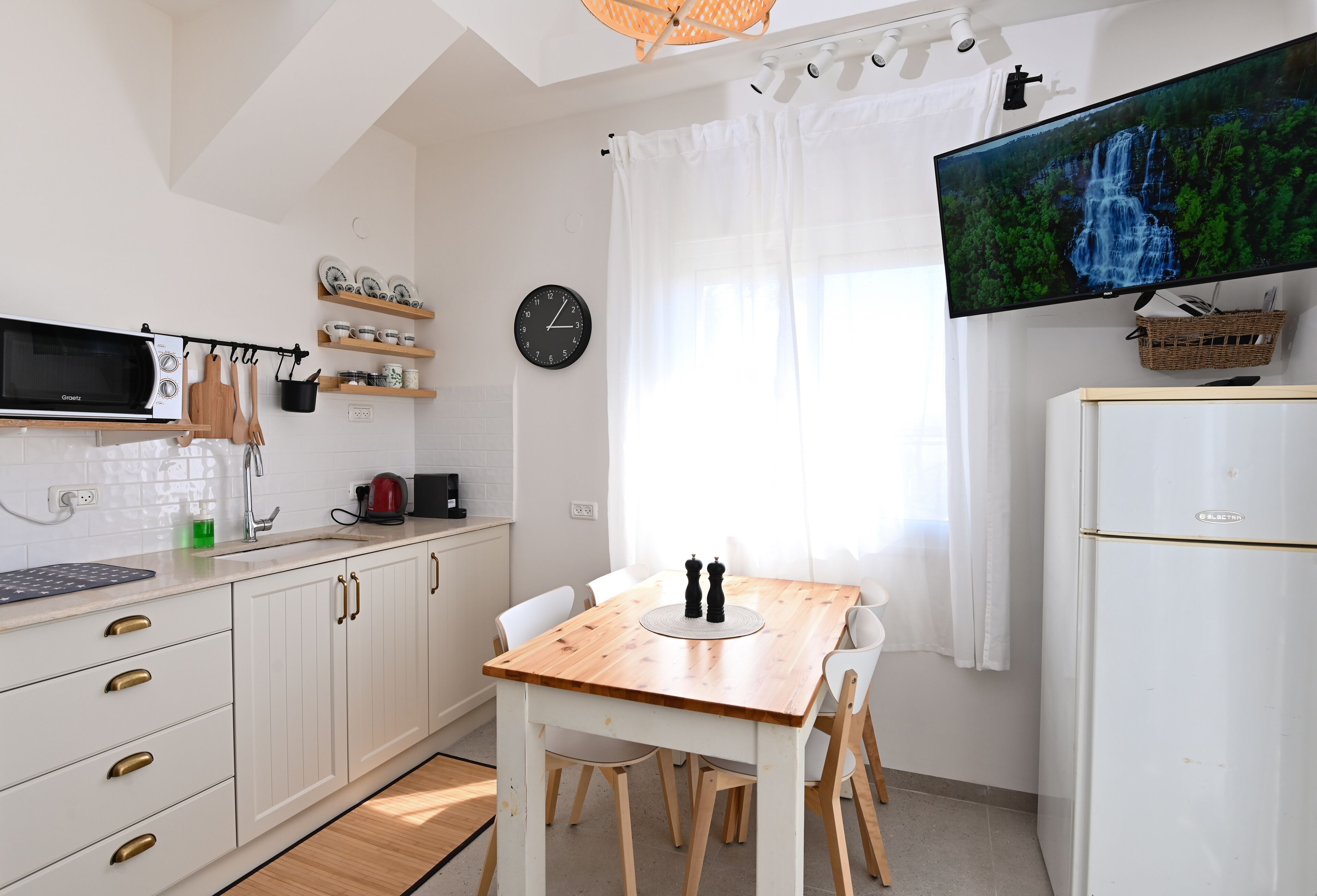 Een moderne keuken met witte kastjes, een houten tafel en een flatscreen televisie waarop een waterval te zien is.