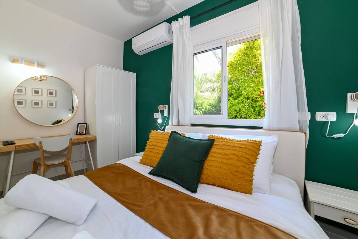 Habitación de hotel con cama, escritorio, silla, armario y ventana con cortinas, y vista a áreas verdes.
