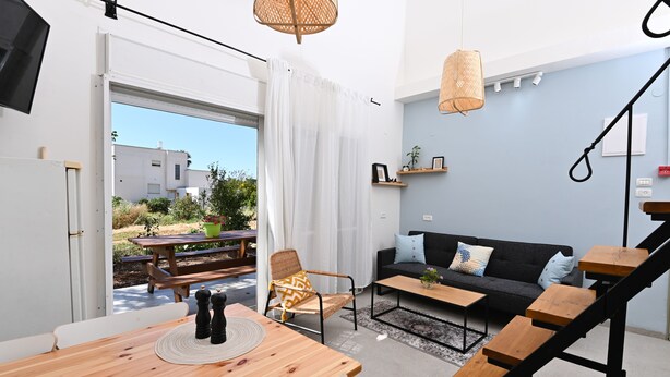 A modern living space with a wooden dining table, a black sofa, and a staircase. - YalaRent Valley view Boutique cottages (Emek Izrael)