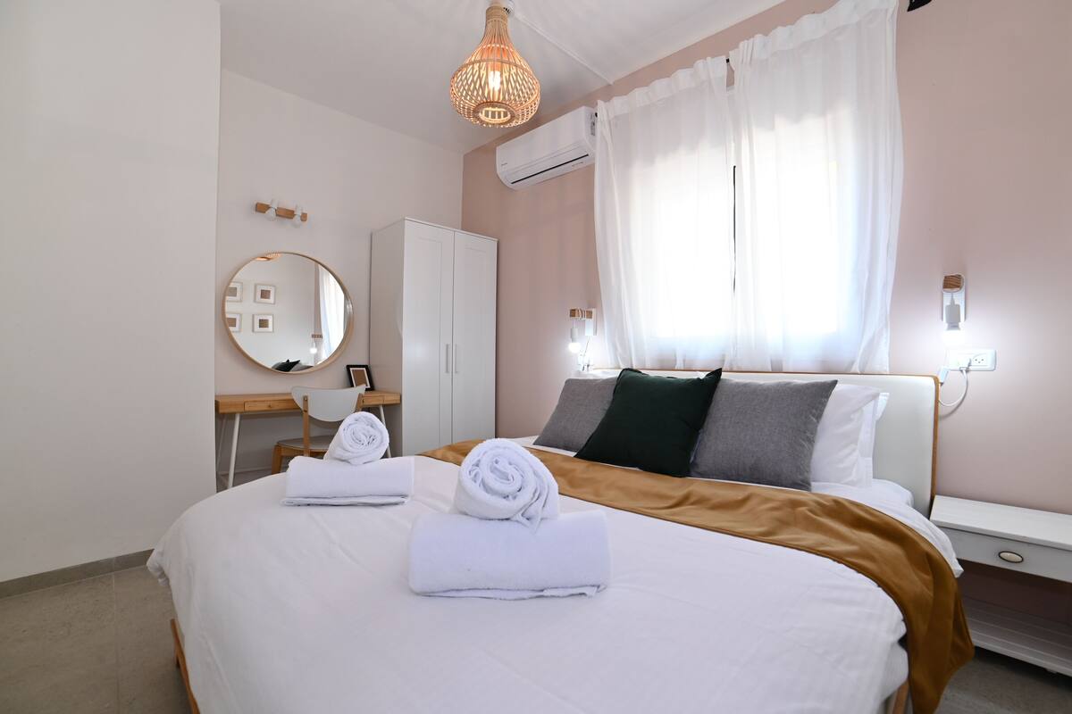 Una cama bien hecha con toallas, una mesita de noche, un espejo y una ventana con cortinas.