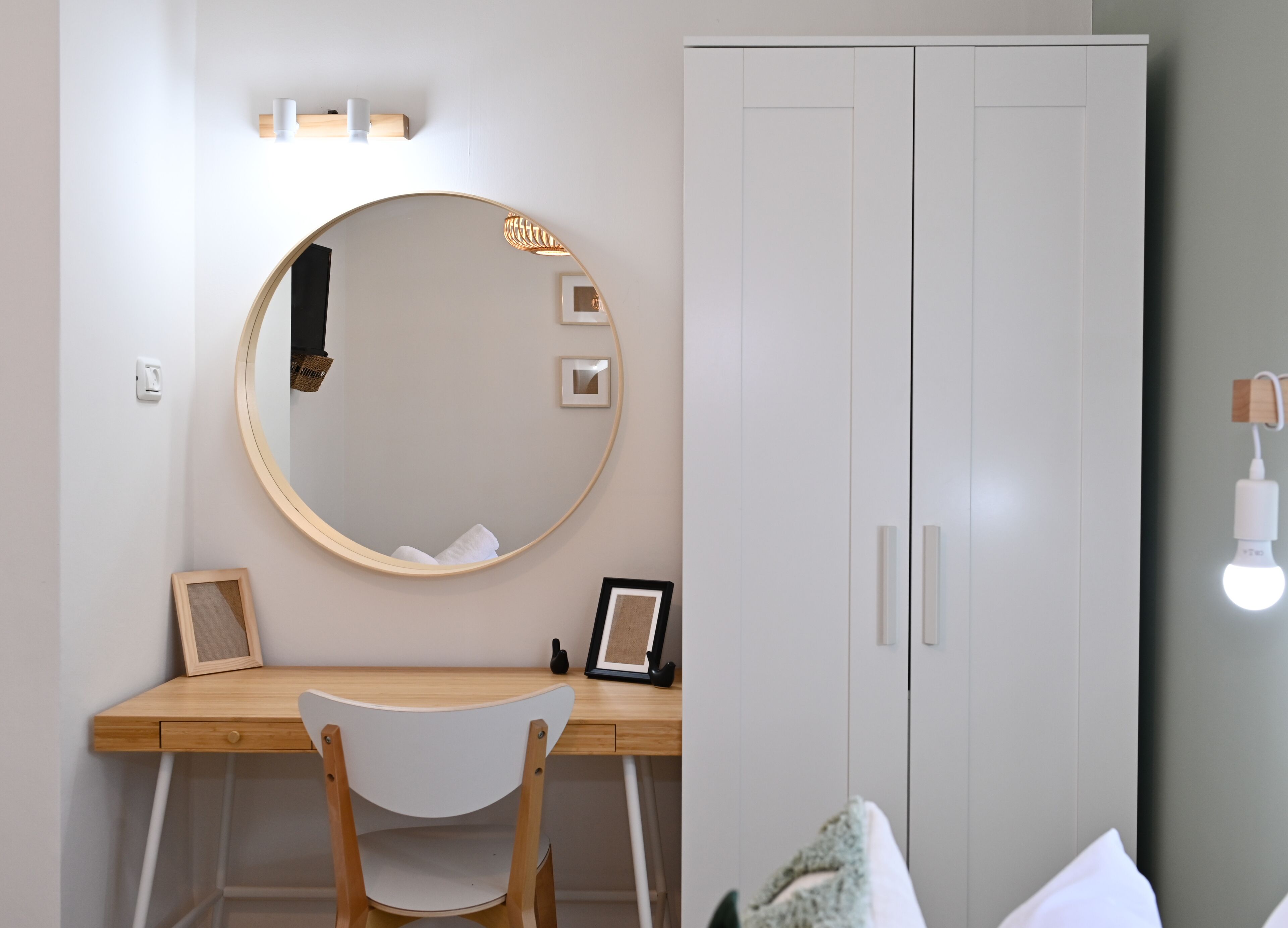 A room with a wooden desk, a round mirror, a wardrobe, and a wall-mounted light fixture.