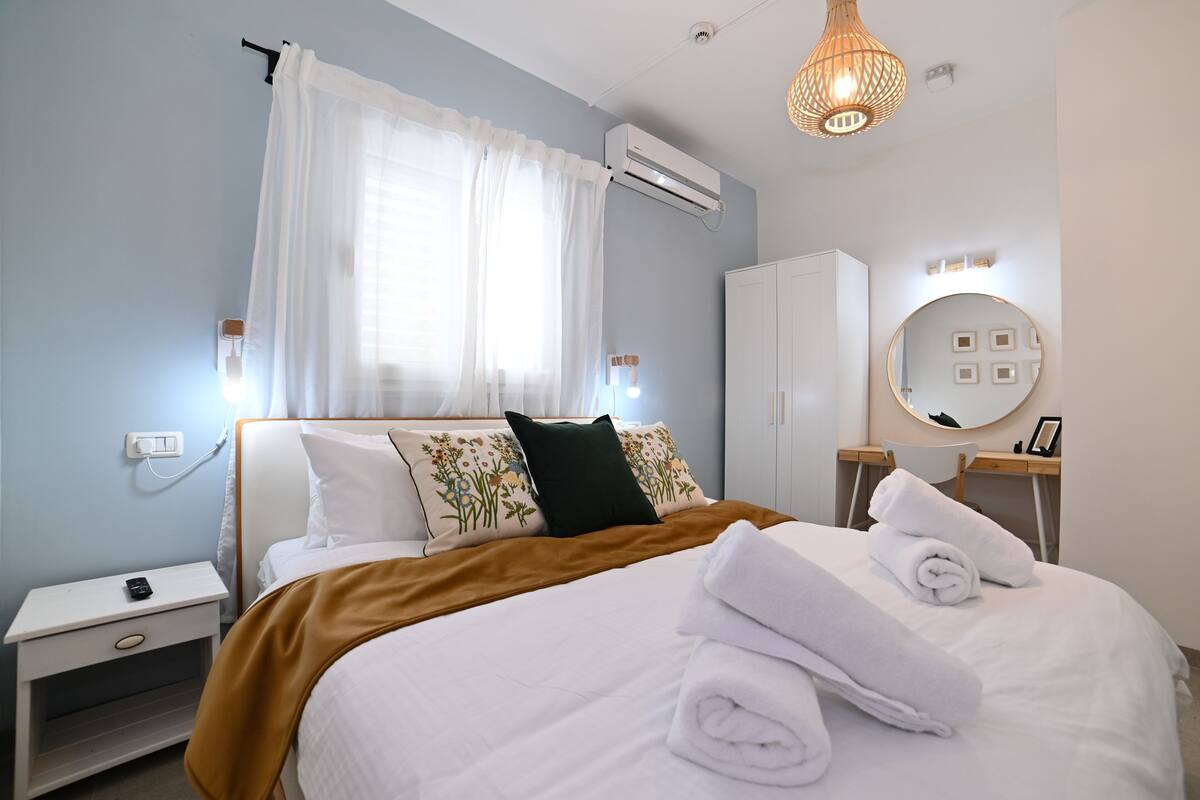 Una cama bien hecha con sábanas blancas y una manta marrón, una mesita de noche con un teléfono, un aire acondicionado instalado en la pared y un espejo redondo con marco cuadrado.