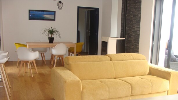 Una sala de estar con un sofá amarillo, una mesa de comedor con sillas, una planta en maceta y un ventanal grande.
