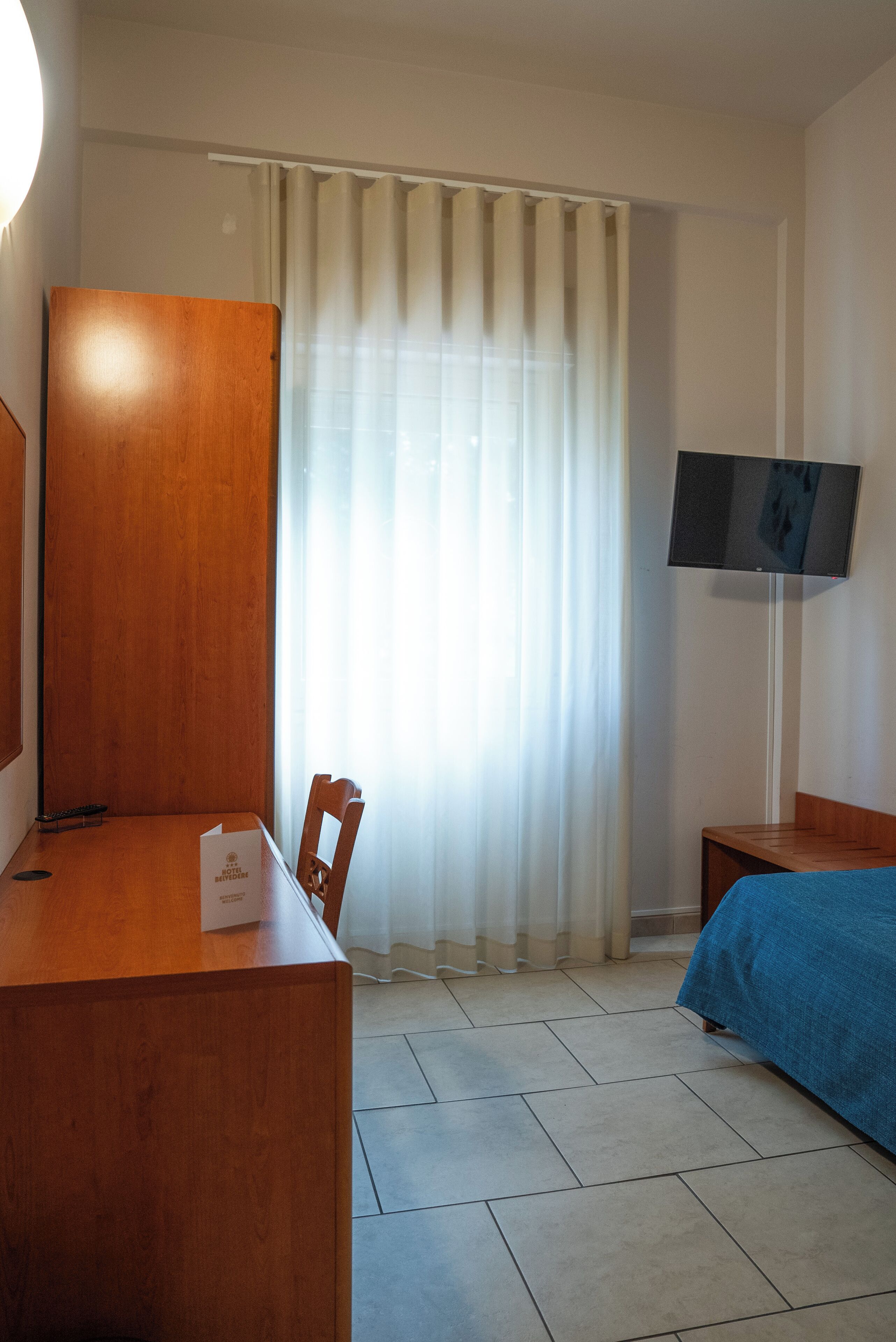 Ett hotellrum med ett skrivbord i trä, en stol, en säng med ett blått överkast, en tv som är monterad på väggen och ett fönster med tunna, genomskinliga gardiner.