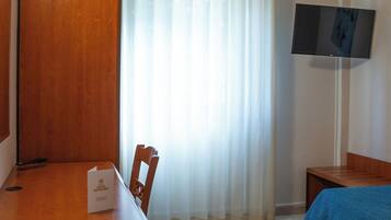 Ett hotellrum med ett skrivbord i trä, en stol, en säng med ett blått överkast, en tv som är monterad på väggen och ett fönster med tunna, genomskinliga gardiner.