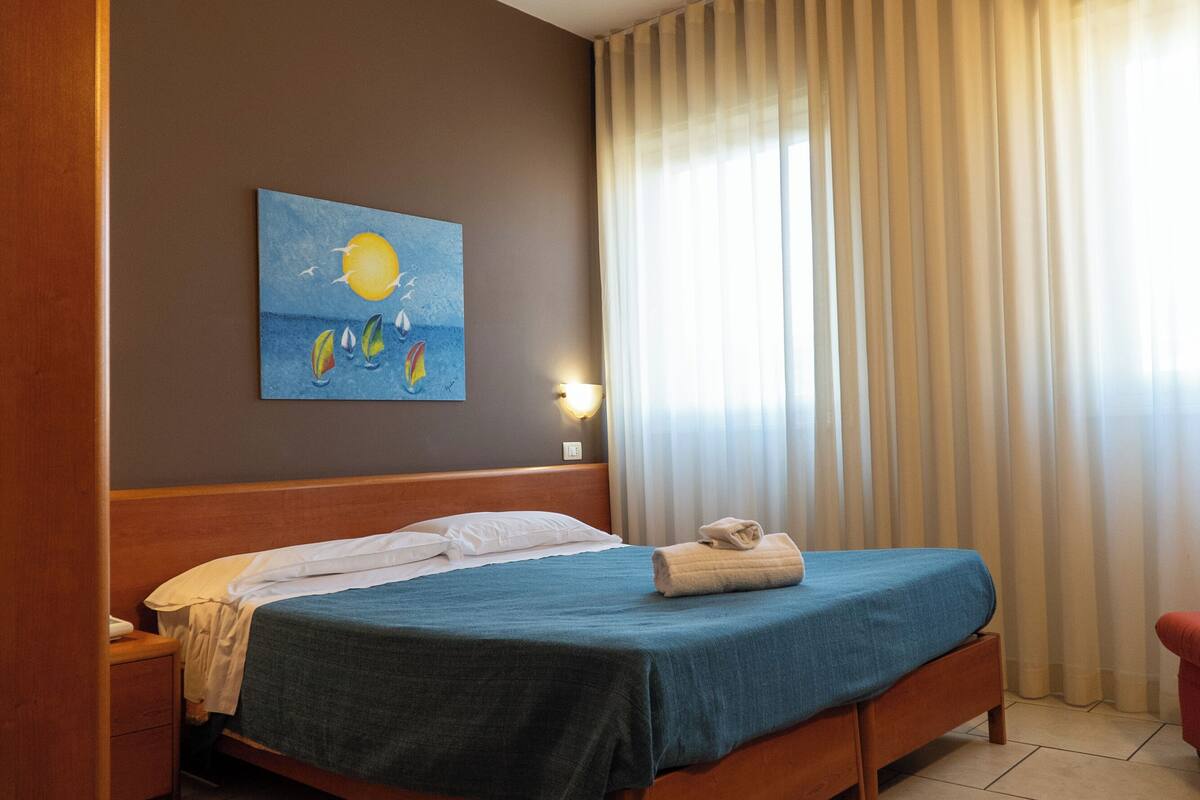 A hotel room with a bed, a painting of a sun and sailboats, a nightstand, and a window with curtains.