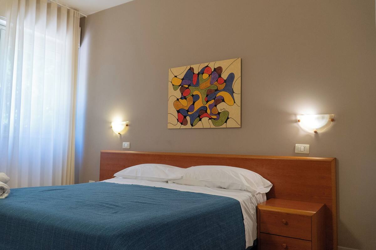 A hotel room with a bed, a wooden nightstand, a wall-mounted abstract painting, and two wall sconces.