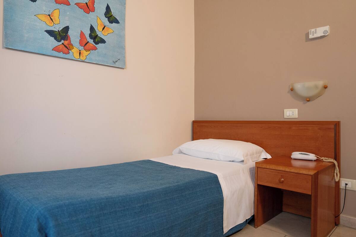 A single bed with a blue bedspread and white pillow, a wooden nightstand with a phone, a wall-mounted light, and a painting of butterflies.