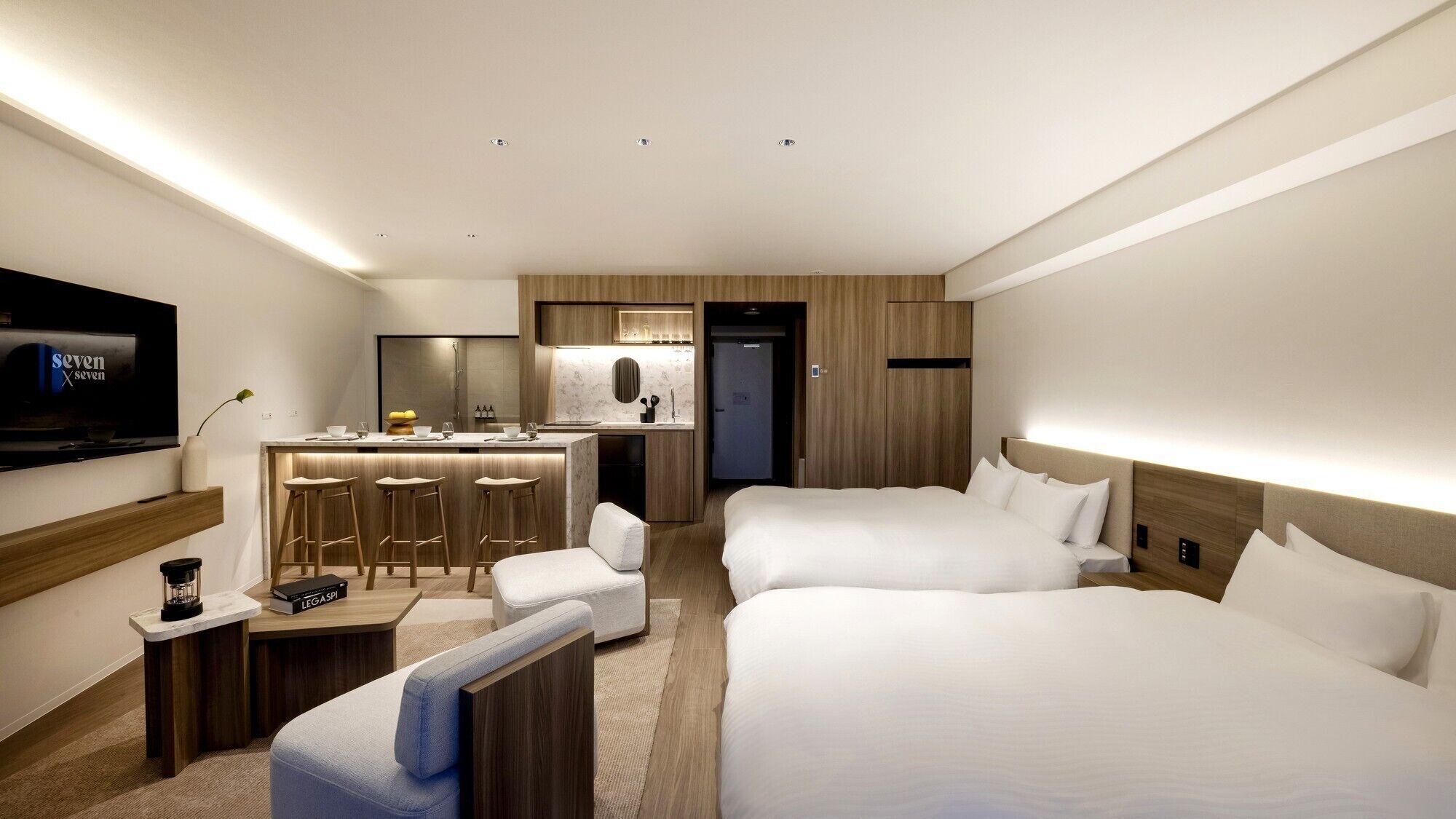 A hotel room with a TV, a kitchenette, two beds, a desk, and a chair.