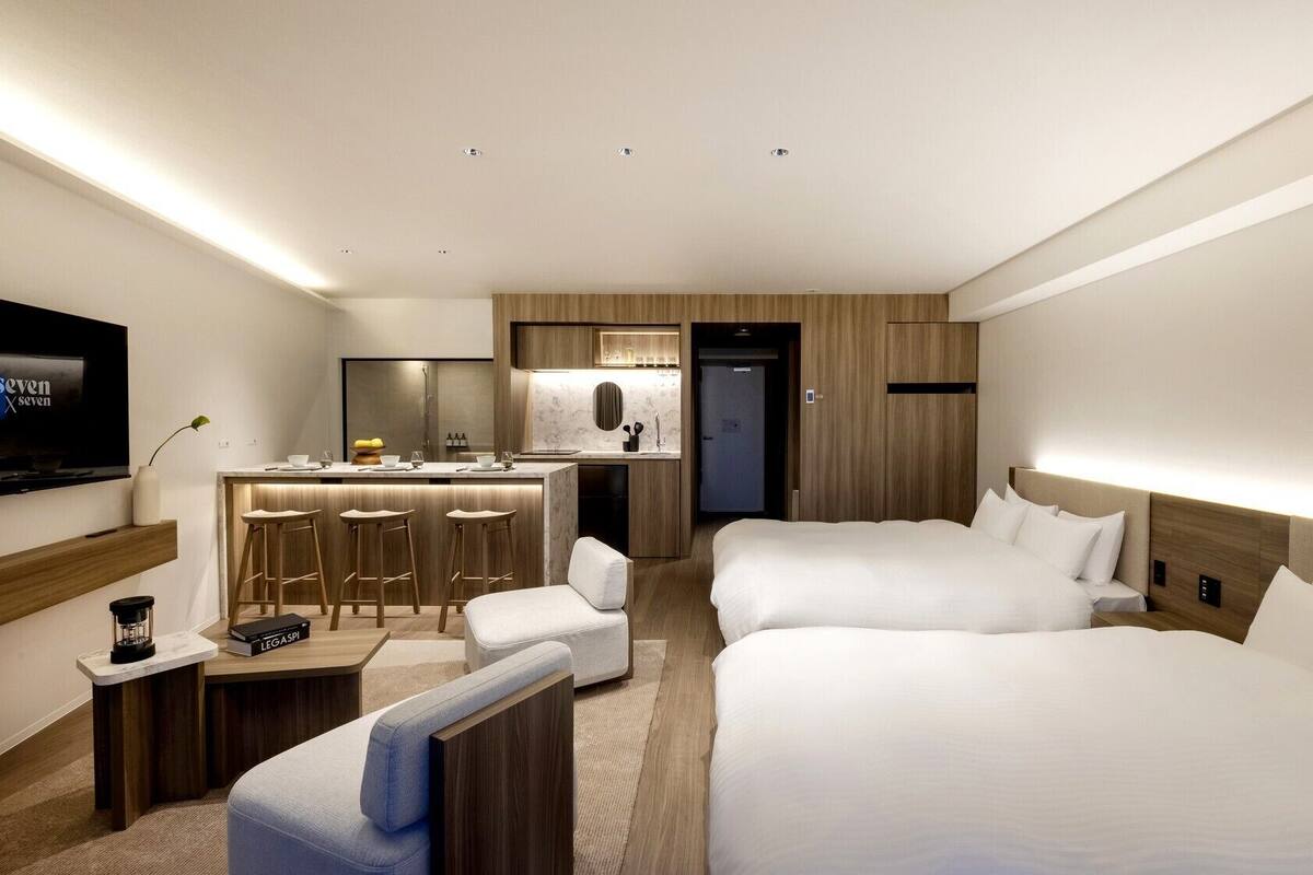 A hotel room with a TV, a kitchenette, two beds, a desk, and a chair.