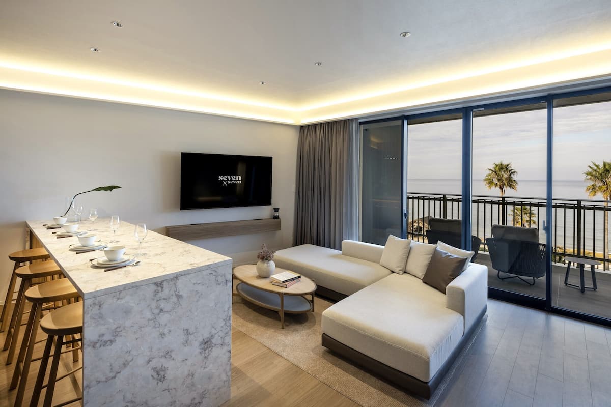 A modern living room with a marble island, a flat-screen TV, a sofa, and a balcony with a view of the sea.