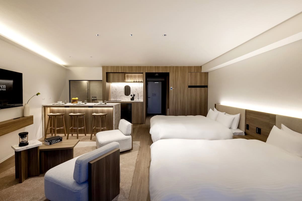 A modern hotel room with a large bed, a smaller bed, a desk with bar stools, a TV, and a kitchenette.