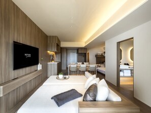 A modern hotel room with a flat-screen TV, a dining area, a kitchen, and a bathroom.