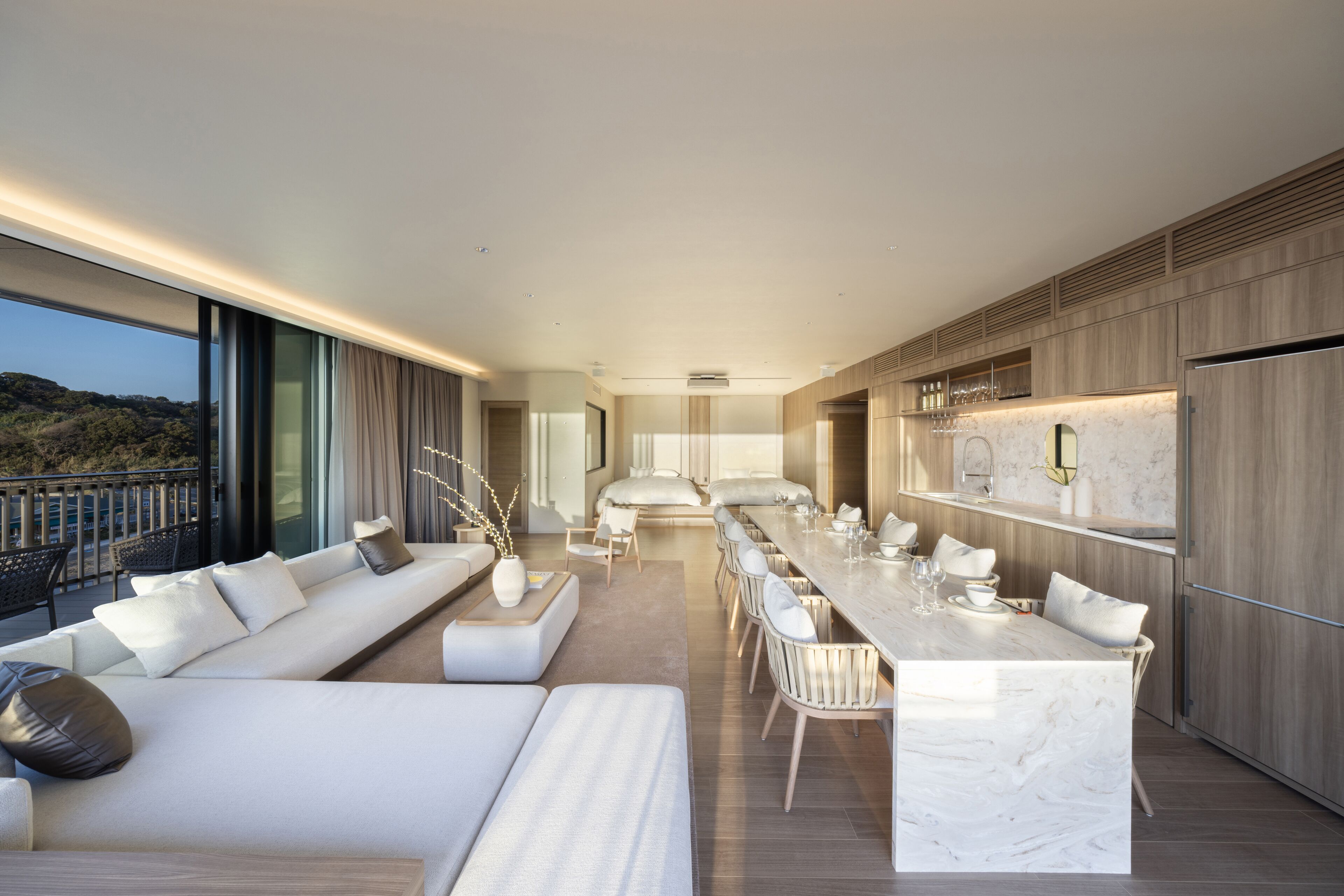 A modern kitchen with a marble countertop and wooden cabinets. There is a dining area with white chairs and a long table. A large window provides a view of the outdoors.