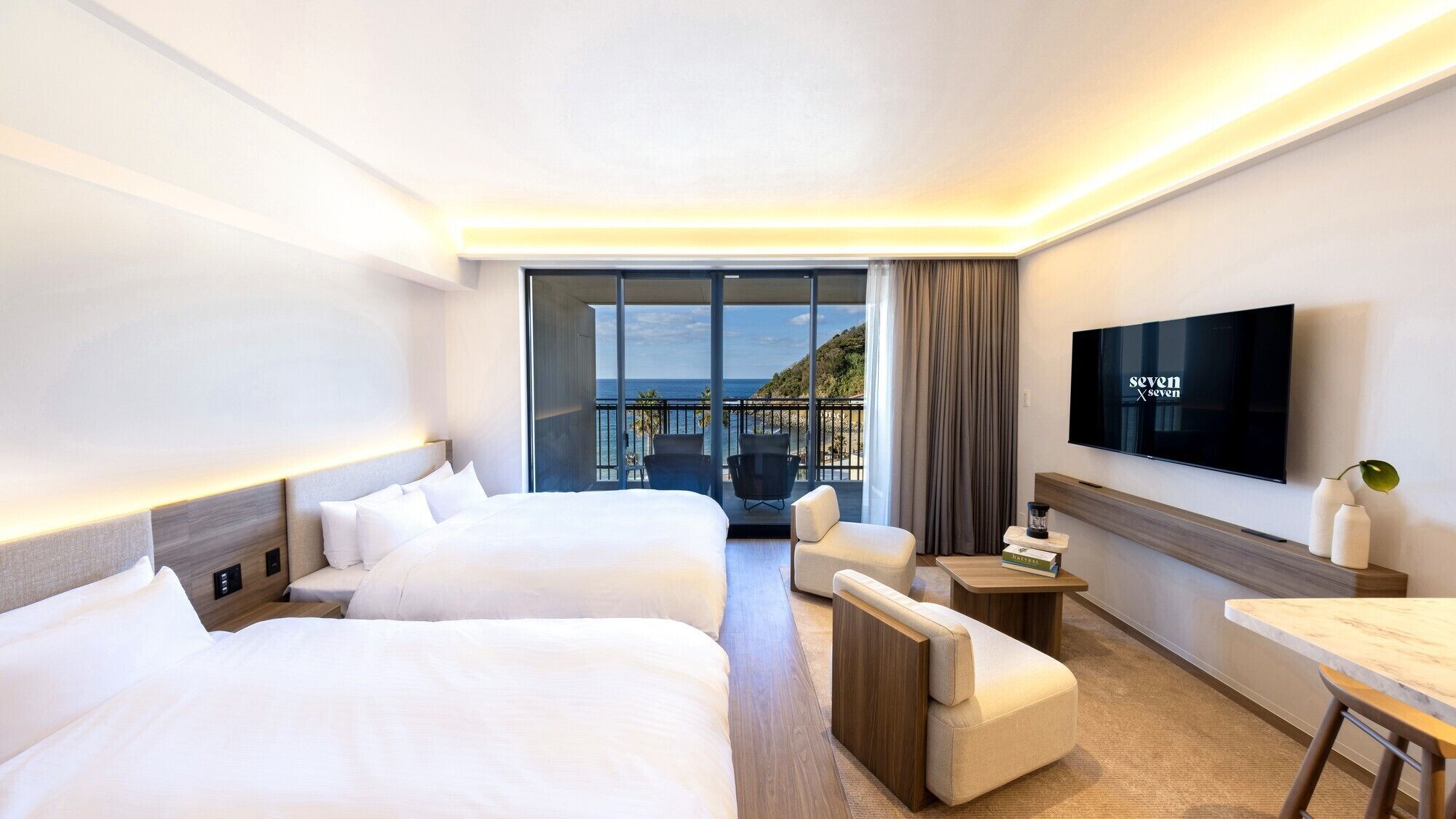 A hotel room with two beds, a TV, a desk, and a view of the sea.