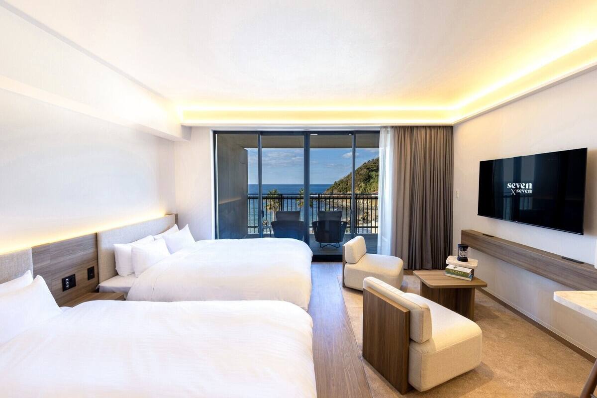 A hotel room with two beds, a TV, a desk, and a view of the sea.