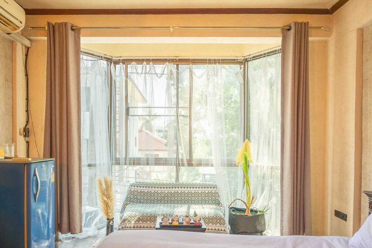 Une chambre avec un lit, une petite table agrémentée d’objets décoratifs, une plante en pot et une fenêtre ornée de rideaux légers.