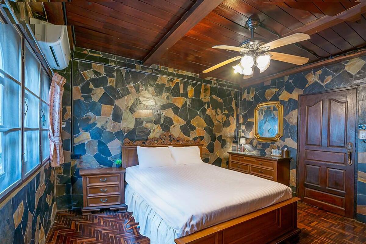 Une chambre avec un mur dont le motif imite la pierre, un lit en bois, un ventilateur de plafond et une porte en bois.