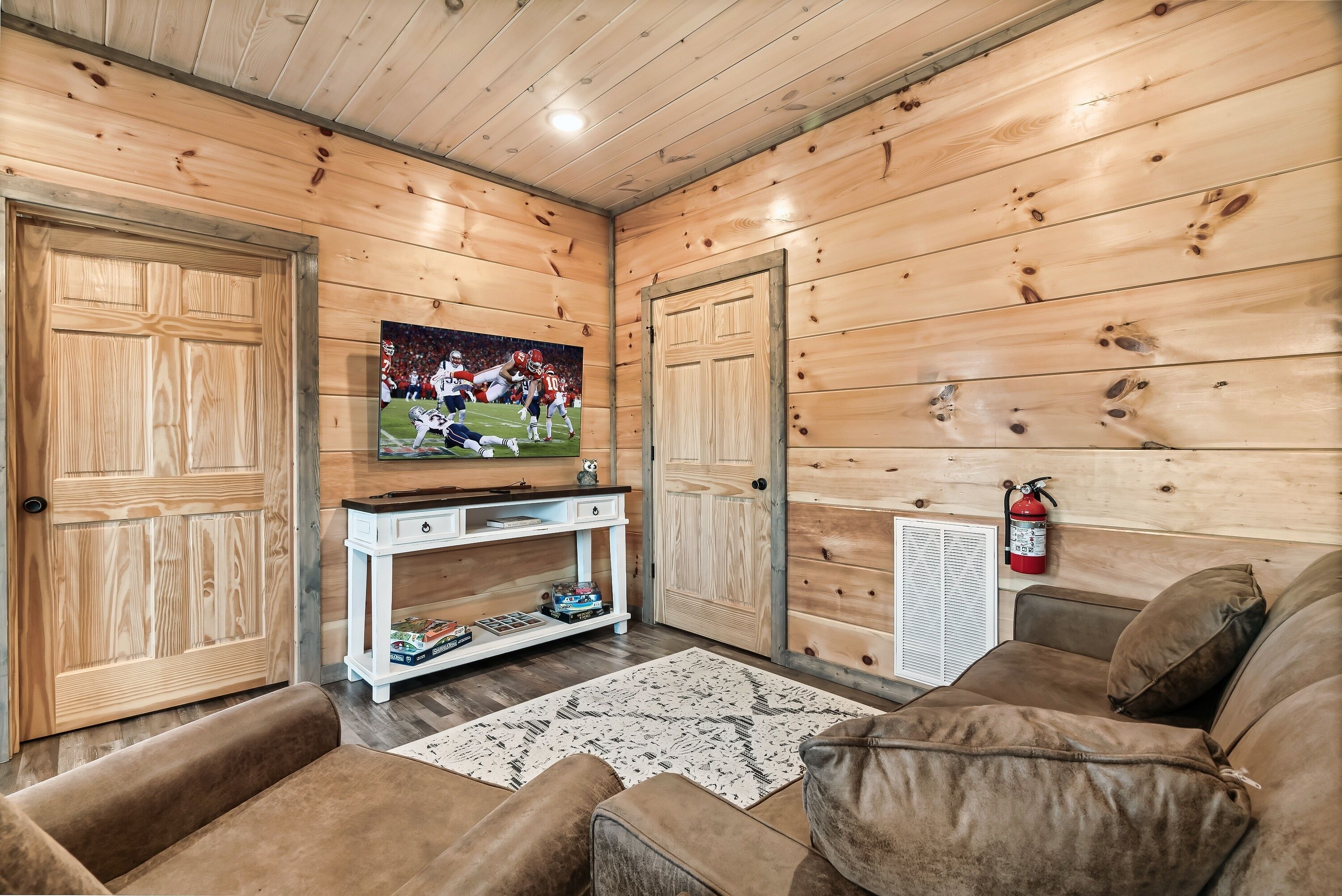 A cozy room with wooden walls, a television mounted on the wall, a white console table, a brown sofa, and a fire extinguisher on the wall.