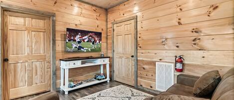 A cozy room with wooden walls, a television mounted on the wall, a white console table, a brown sofa, and a fire extinguisher on the wall.
