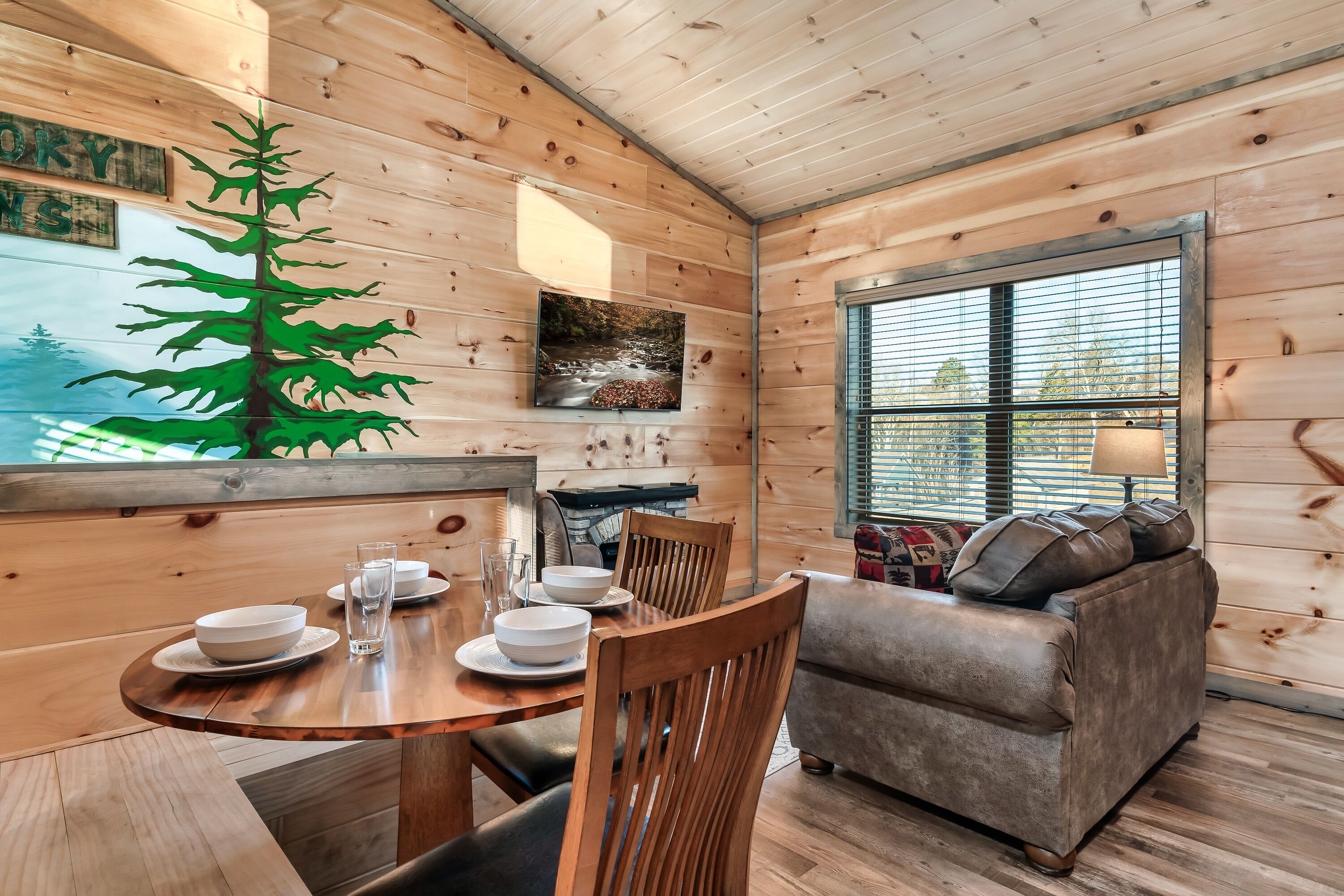 A wooden cabin interior with a dining table set for four, a sofa, and a large window.