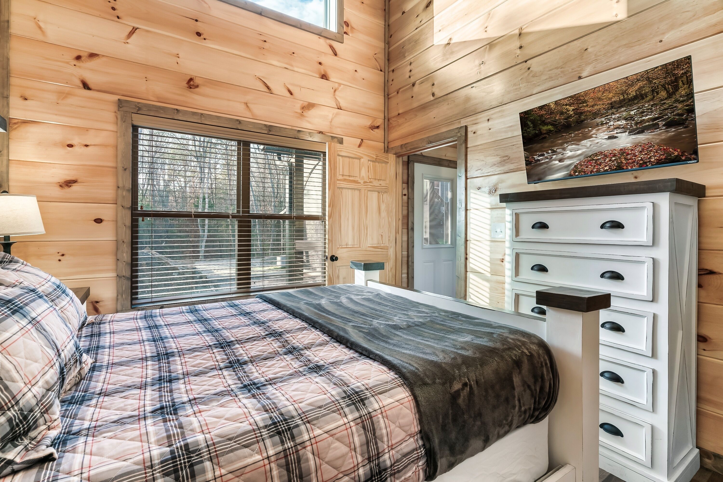 A bedroom with a plaid bedspread, a wooden ceiling, a window with blinds, a dresser with drawers, and a painting above the dresser.