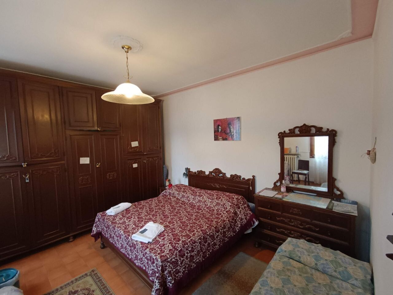 A bedroom with a wooden wardrobe, a bed with a patterned bedspread, a dresser with a mirror, and a small rug on the floor.