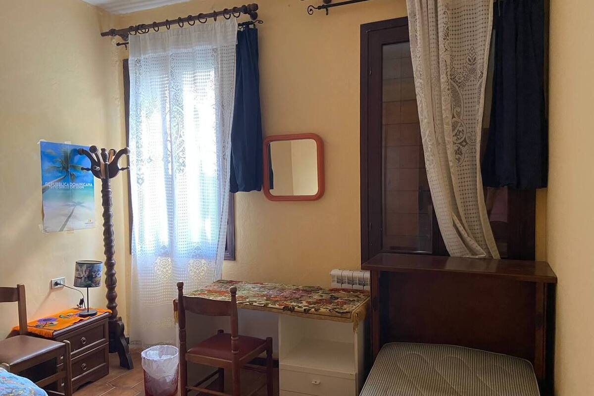 A small, well-lit bedroom with a single bed, a desk with a chair, a mirror, and a window with curtains.