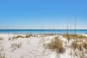 Une plage de sable avec quelques touffes d’herbe, un ciel dégagé et une mer calme.