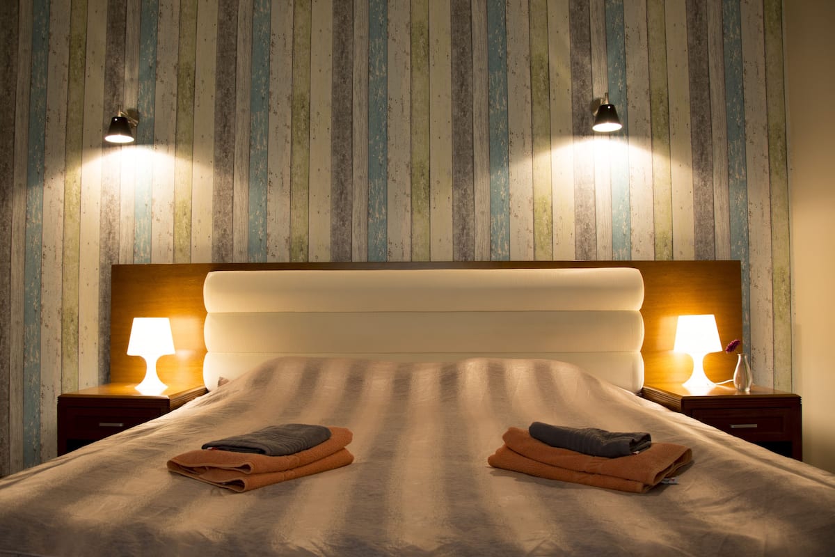 Ein ordentlich bezogenes Bett mit zwei gefalteten Handtüchern, zwei Nachttischlampen und einem Holz-Kopfteil.