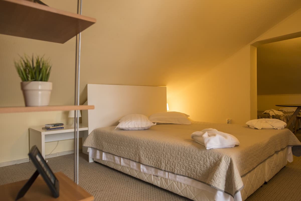 Ein ordentlich bezogenes Bett mit Kissen und Decke, ein Nachttisch mit Lampe und Pflanze sowie ein kleines Regal mit Büchern.