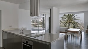 Una cocina moderna con una isla de acero inoxidable, horno integrado y un amplio ventanal con vistas a palmeras y un paisaje urbano.
