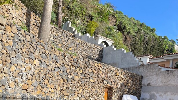 Un muro de piedra con una pequeña ventana, una piscina cristalina que refleja el cielo y una palmera.