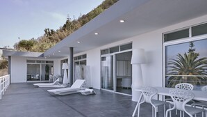 Un área lounge moderna al aire libre con mobiliario blanco, incluyendo sillas y una mesa, y un ventanal que da a un espacio interior.
