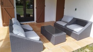 Eine Terrassenfläche mit Korbmöbeln, darunter zwei Sofas und ein Couchtisch, sowie eine Glastür, die in einen Garten führt.